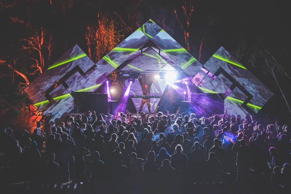 Optimus Gryme playing to a packed bush doof crowd, the stage lit green and purple under the trees