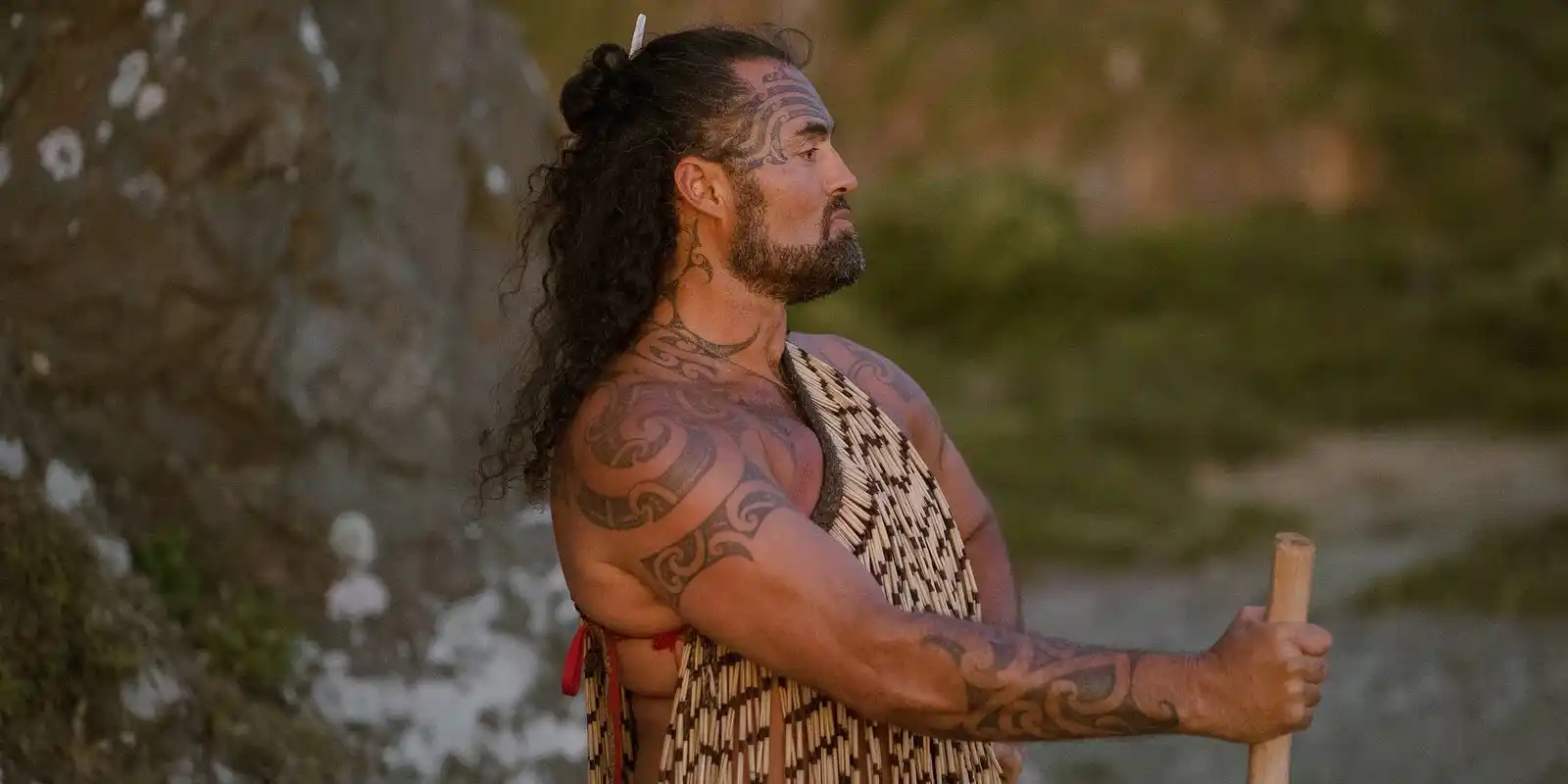 Matiu Te Huki in korowai, standing with a taiaha beside the water
