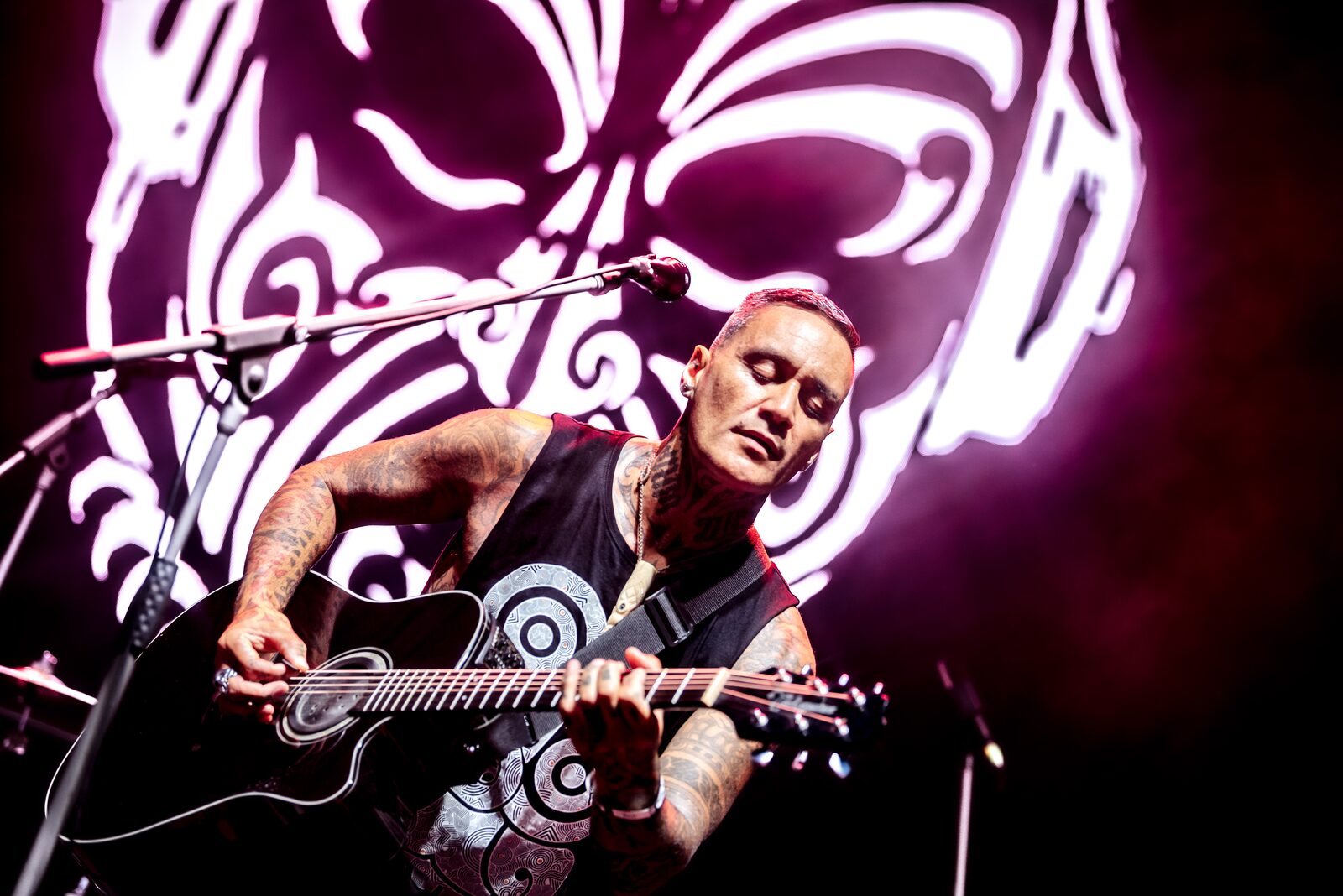 Tiki Taane performing on stage with a guitar, lit by projected moko