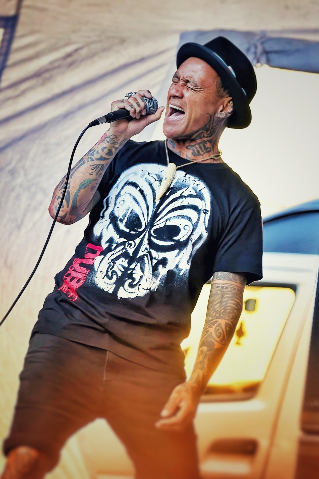 Tiki Taane singing into a handheld microphone on the main stage, wearing a black hat and a Māui-print tee